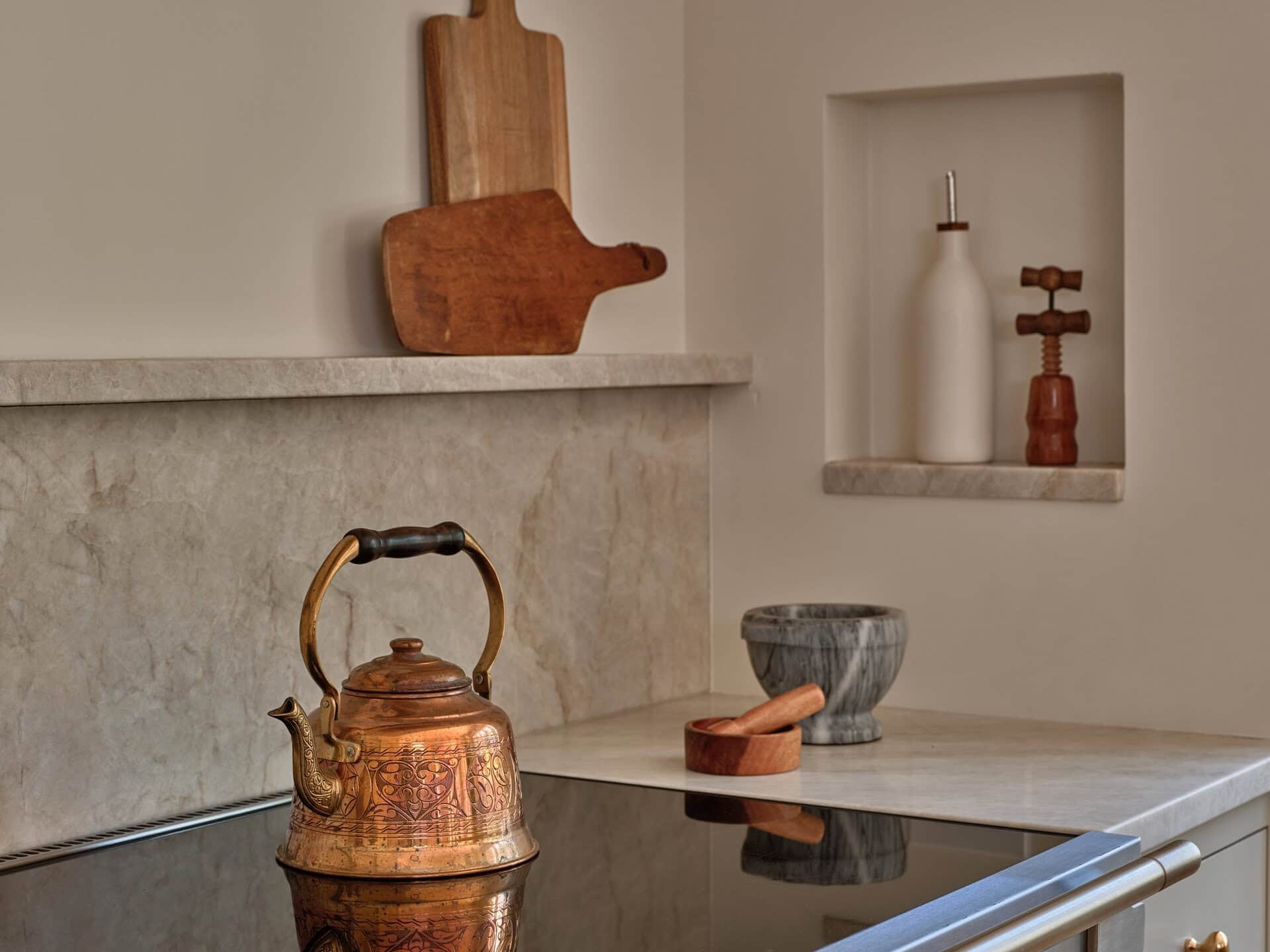 A copper tea kettle sits on a black glass stovetop in front of a stone backsplash with a wall niche containing a bottle and a corkscrew.