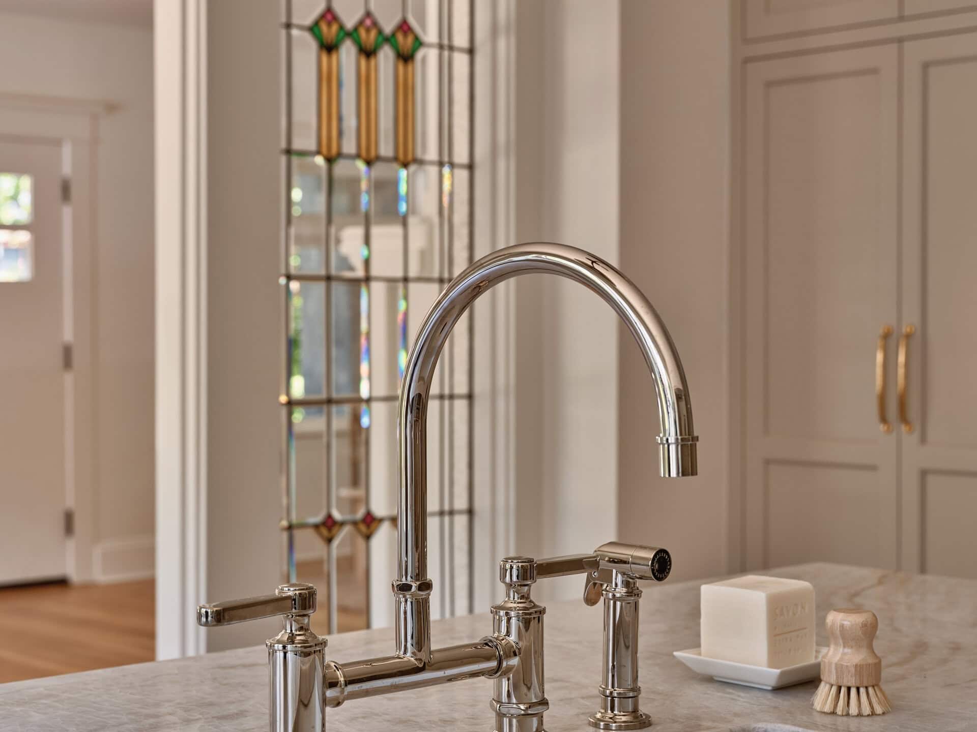 Polished nickel bridge kitchen faucet mounted on a marble countertop next to a bar of soap and a wooden scrubbing brush.